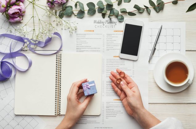 Wedding background with checklist and calendar. Female hands holding rings and arranging marriage, filling in planners on white wooden table with lots of tender bridal stuff, top view